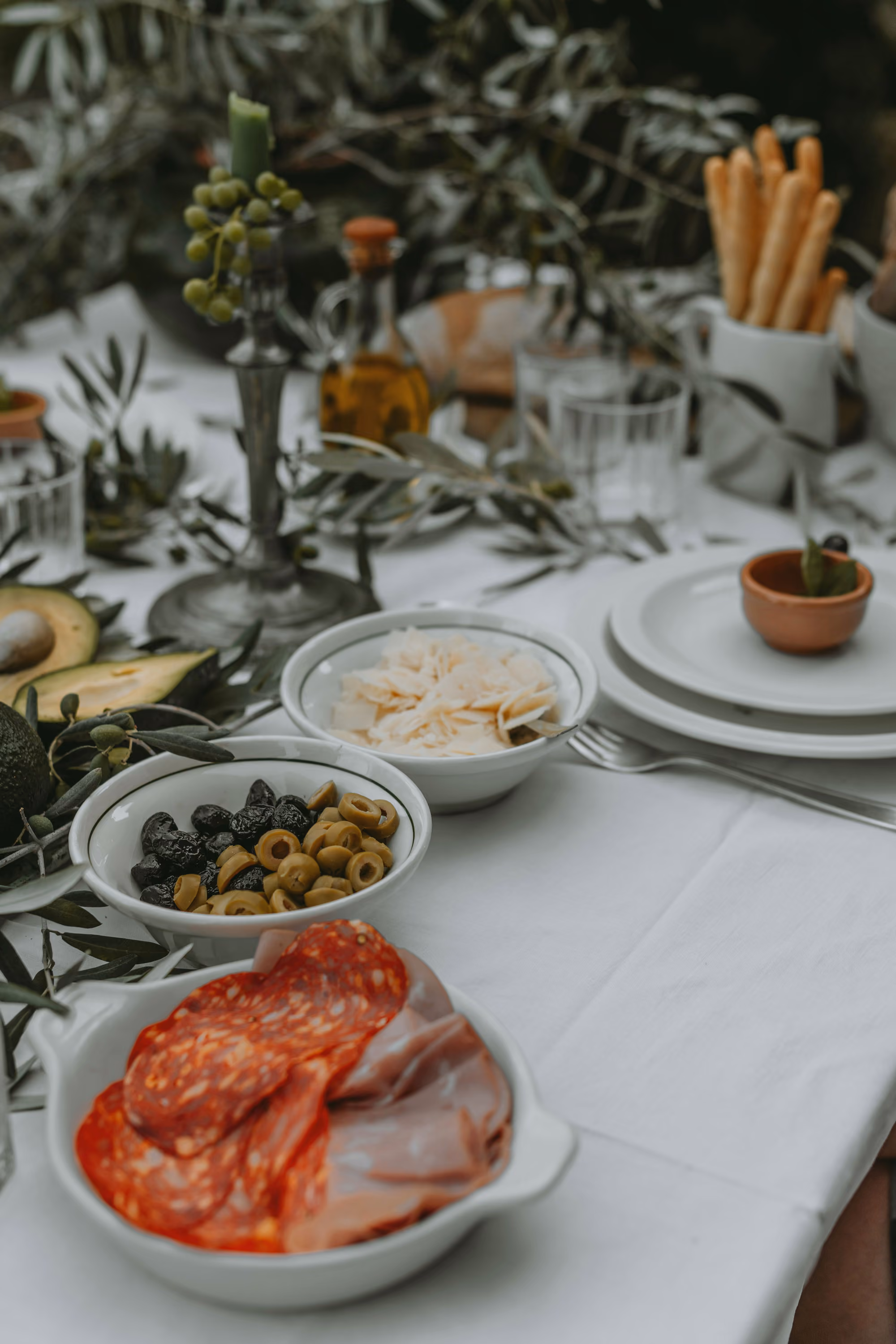 Table set with white tablecloth featuring plates, glassware, and bowls of sliced cured meats, black and green olives, shaved cheese, breadsticks in a container, avocado halves, olive branches, and a candle holder with an olive branch decoration.