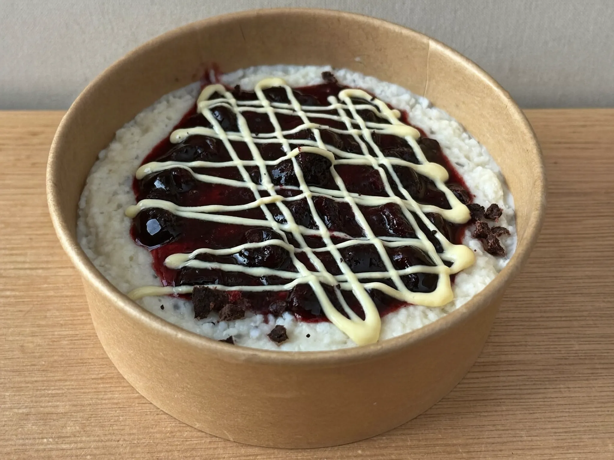 Milchreis-Bowl in brauner Pappschale mit roter Grütze, Vanillesauce-Linien und Oreo-Crumble