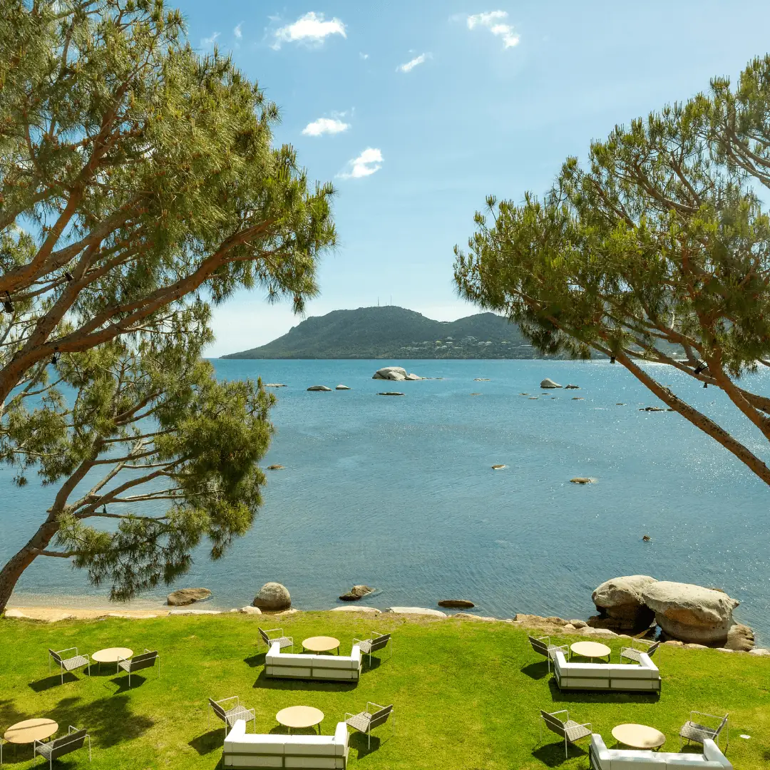 Vue sur une baie calme avec un îlot rocheux au milieu de l'eau bleue, encadrée par deux grands pins, et un espace détente extérieur avec des canapés blancs et des tables rondes sur une pelouse verte.