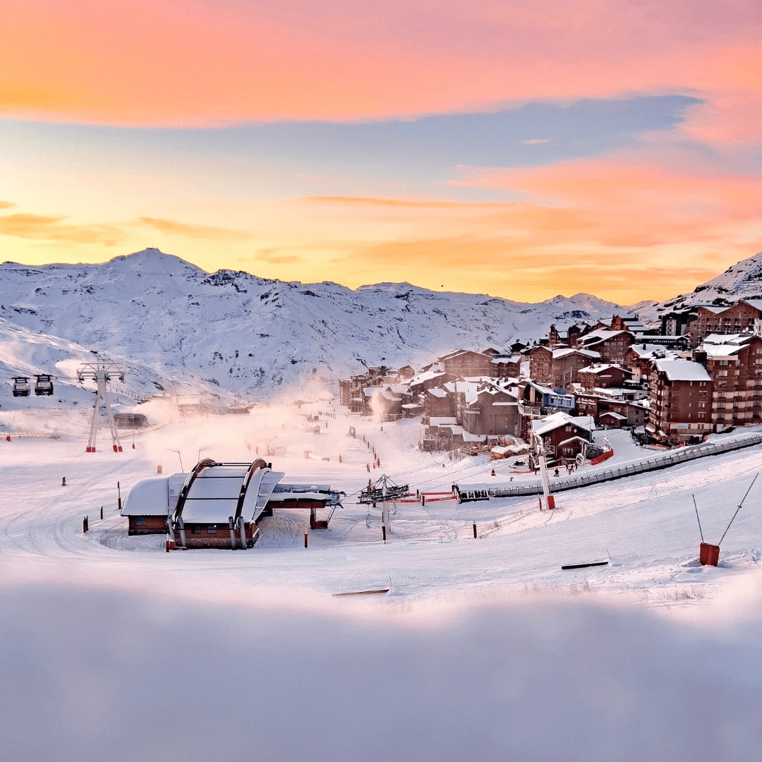 Station de ski avec remontées mécaniques et chalets enneigés au pied de montagnes couvertes de neige sous un ciel au coucher du soleil aux teintes rose et orange.