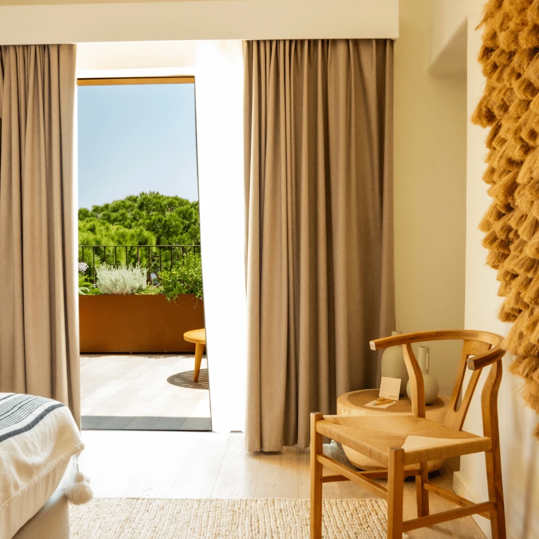 Coin d'une chambre lumineuse avec un lit à gauche, une chaise en bois clair à droite, une table ronde avec un vase, et une porte ouverte donnant sur une terrasse avec des plantes vertes et vue sur des arbres sous un ciel bleu.