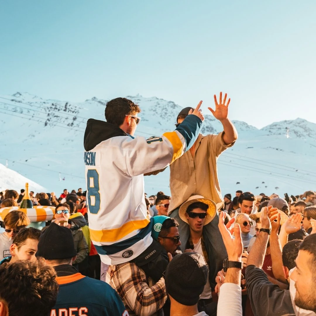 Un grand groupe de personnes danse et célèbre en plein air dans une zone montagneuse enneigée, certaines portant des lunettes de soleil et des chapeaux, avec un ciel clair en arrière-plan.