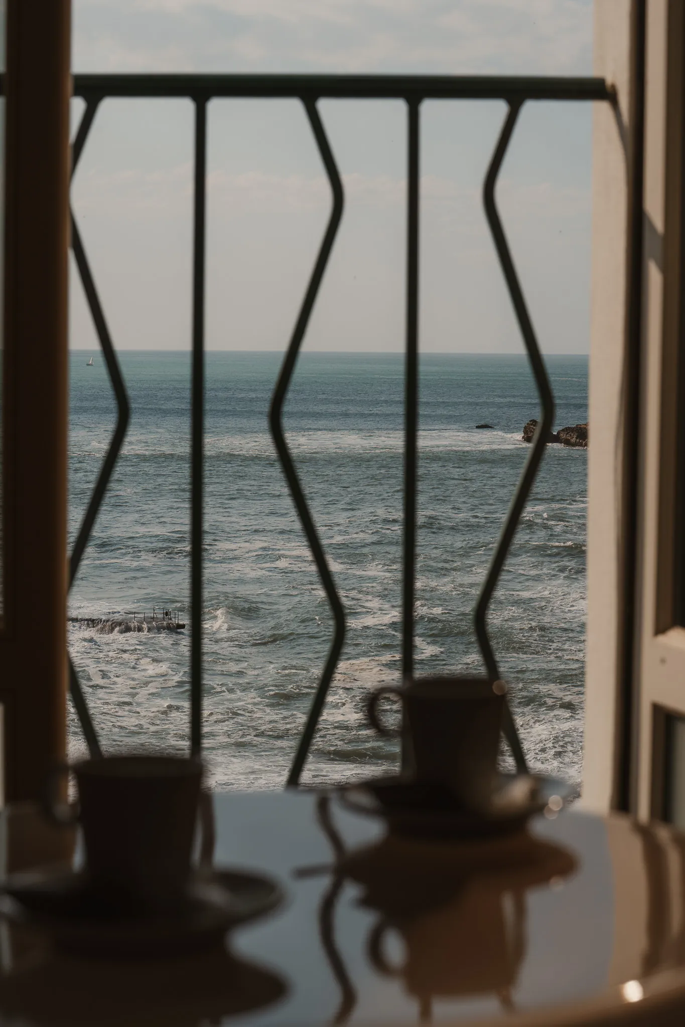 Vue sur l'océan depuis un balcon avec une balustrade en métal, une table avec deux tasses à café floues au premier plan.