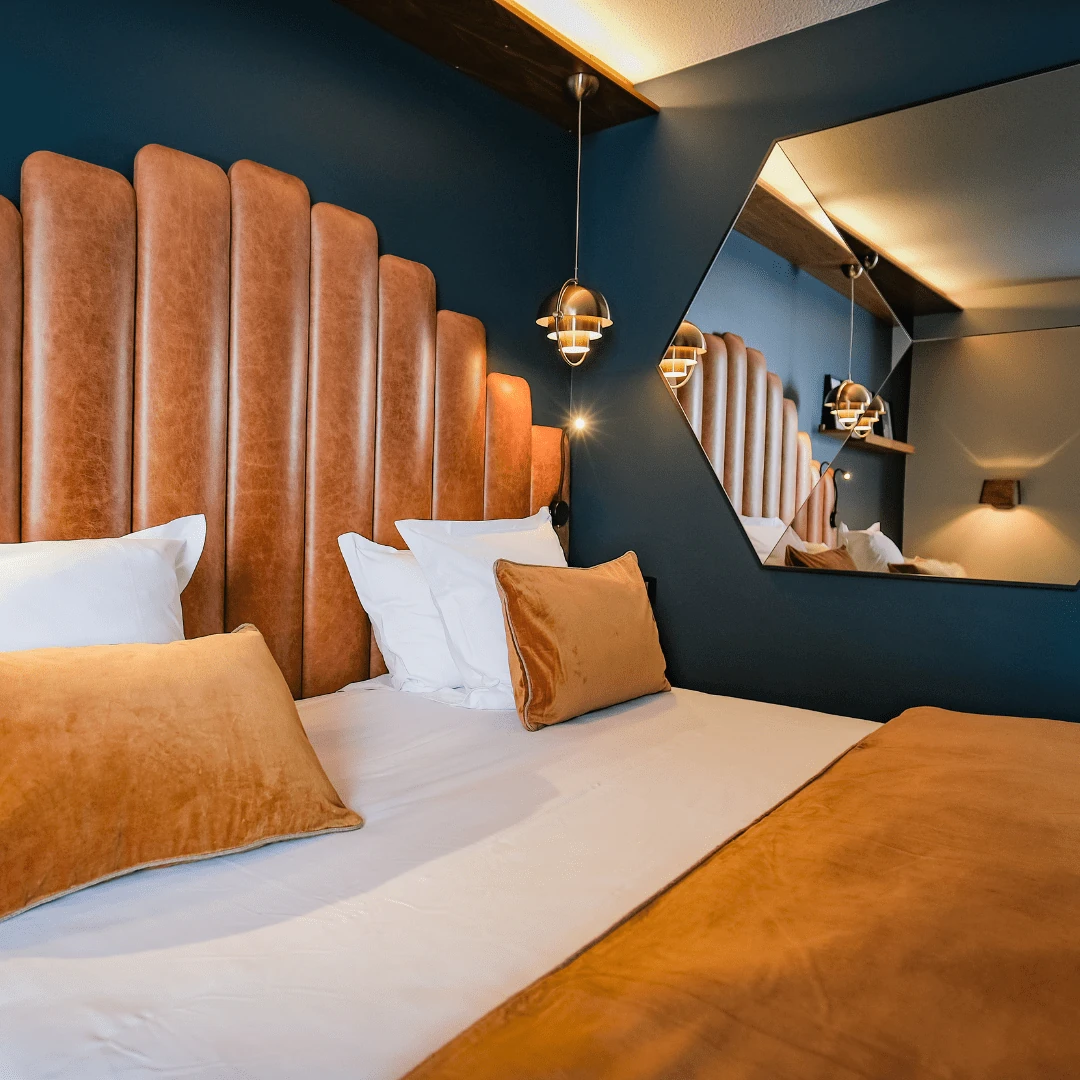 Chambre moderne avec grand lit, tête de lit en simili cuir brun, oreillers blancs et coussins marron, mur bleu foncé, miroir hexagonal réfléchissant la pièce et lampe suspendue sombre.
