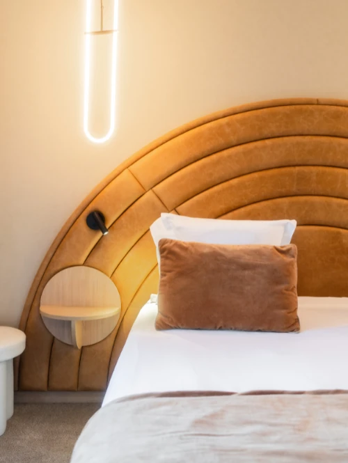 Une partie d'un lit avec tête de lit incurvée en cuir brun, coussin marron et un lampadaire vertical lumineux fixé au mur clair à gauche.