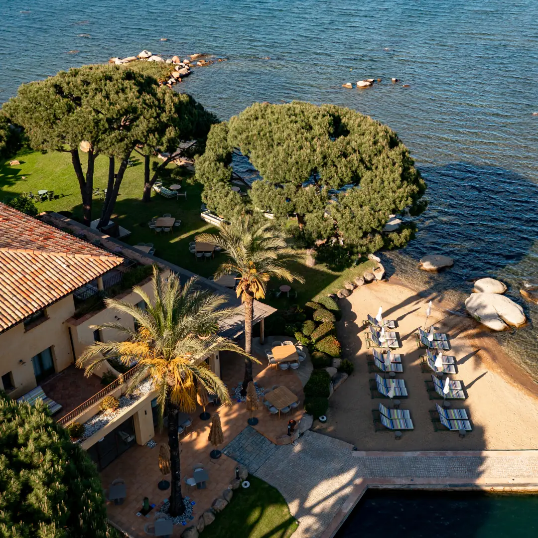 Vue aérienne d'une zone de villégiature au bord de l'eau avec des arbres, des tables de jardin, des chaises longues sur une petite plage de sable et une piscine.