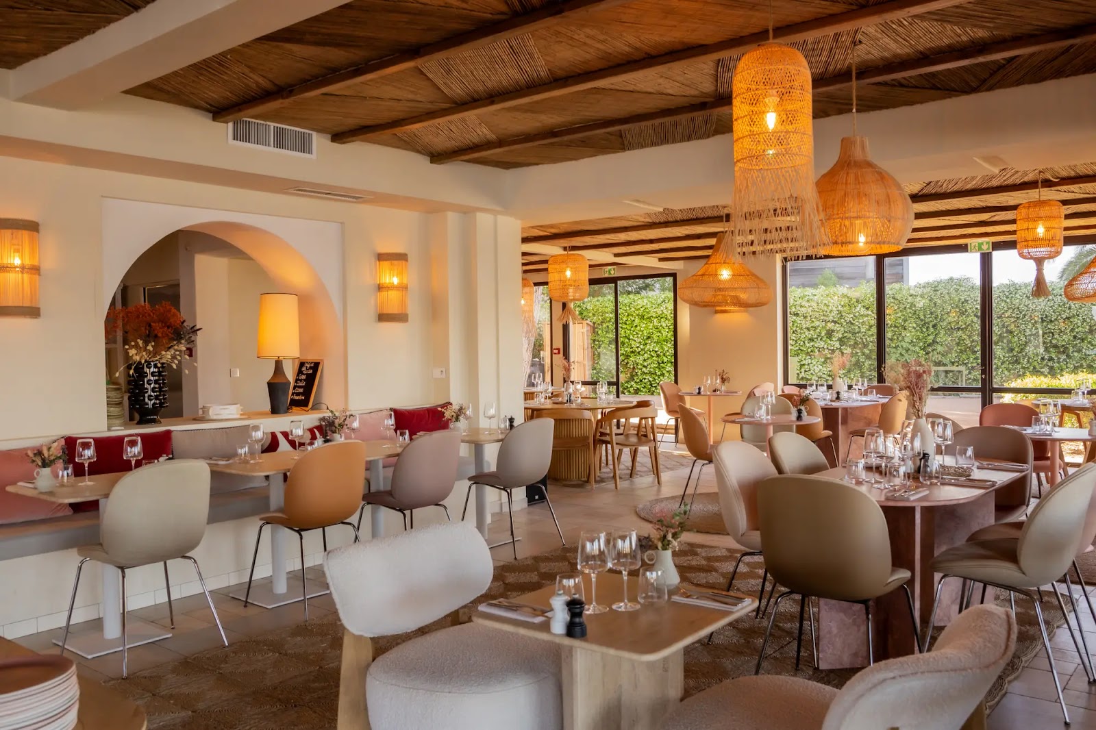 Intérieur d'un restaurant moderne avec tables en bois, chaises de différentes couleurs, suspensions en osier et fenêtres donnant sur un jardin vert.