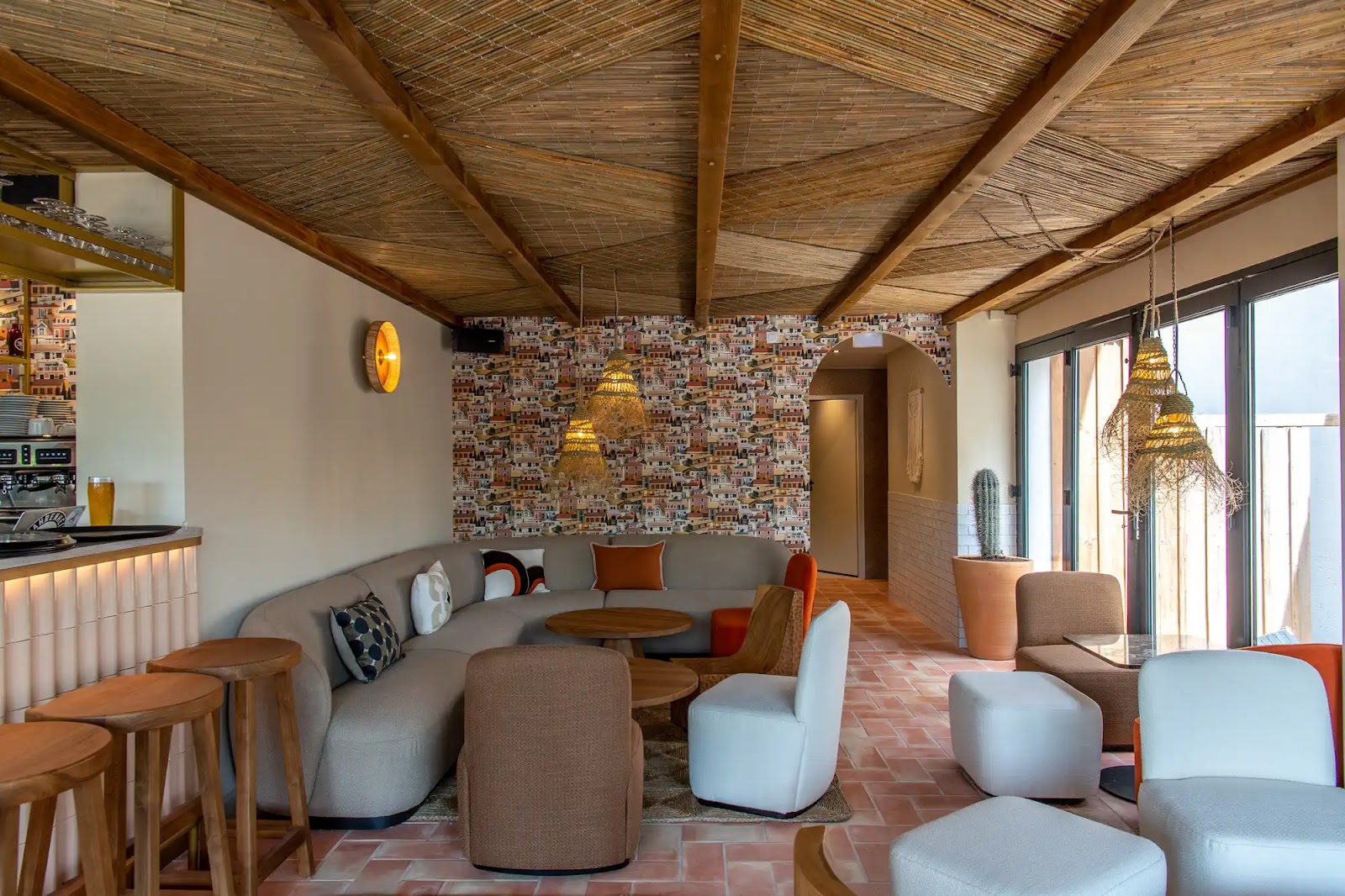 Salon intérieur avec canapé incurvé gris, plusieurs fauteuils de couleurs neutres, tables basses en bois, mur avec papier peint de maisons colorées, plafonds en bambou, luminaires suspendus en corde, grandes fenêtres vitrées et sol en carreaux terre cuite, décor chaleureux et convivial.