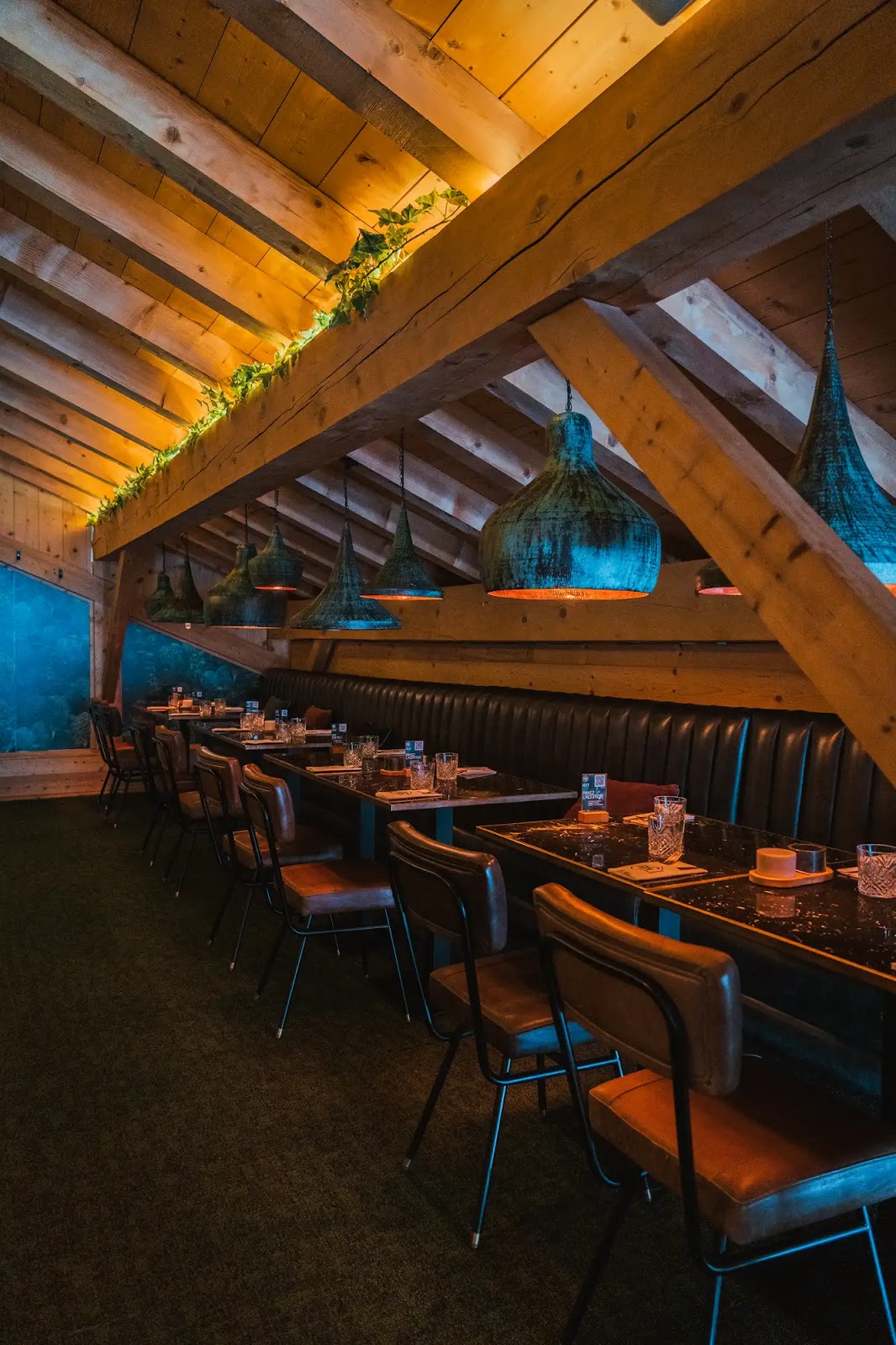 Intérieur chaleureux d'un restaurant avec plafond en bois à poutres apparentes, rangée de tables en marbre noir, chaises en cuir marron et banquette en cuir noir, éclairage doux par des suspensions vintage en métal patiné, et décoration végétale au-dessus d'une poutre.