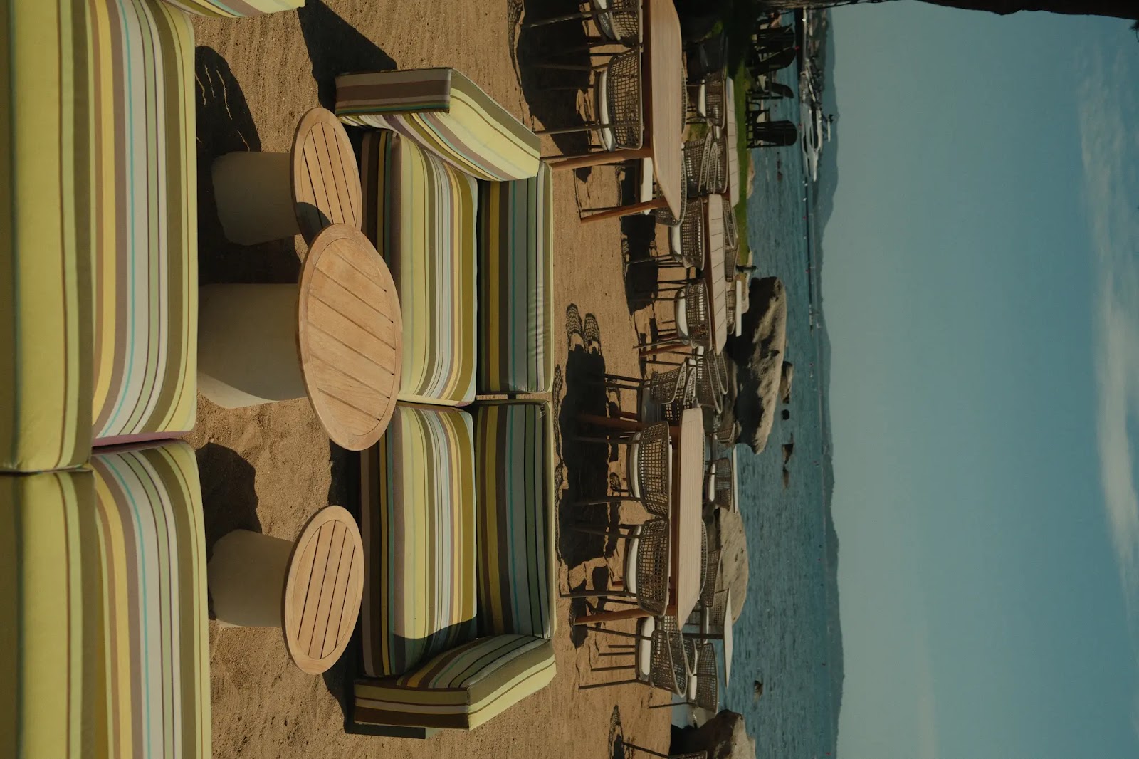Chaises longues et tables en bois avec coussins rayés verts, jaunes et blancs sur une plage sablonneuse devant un lac avec des rochers et des bateaux à l'horizon sous un ciel bleu.