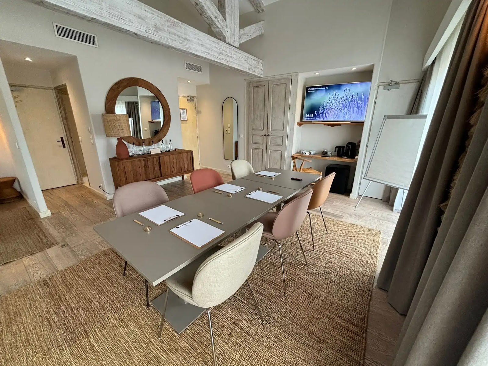 Salle de conférence moderne avec une table grise entourée de six chaises pastel, chaque place équipée d'un bloc-notes, d'un stylo et d'une pince, moquette tissée, murs clairs avec poutres apparentes blanches, grand miroir rond au-dessus d'un meuble en bois, télévision à écran plat affichant une image de lavande, tableau blanc sur pied près des rideaux.
