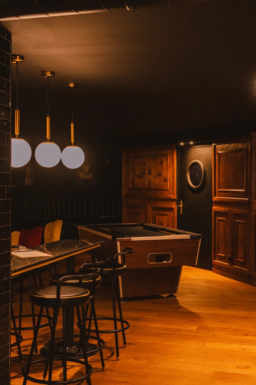 Intérieur chaleureux d'une salle de loisirs avec table de billard, tabourets noirs autour d'une table à plateau en marbre, suspensions lumineuses sphériques et meubles en bois foncé sur un sol en bois clair.