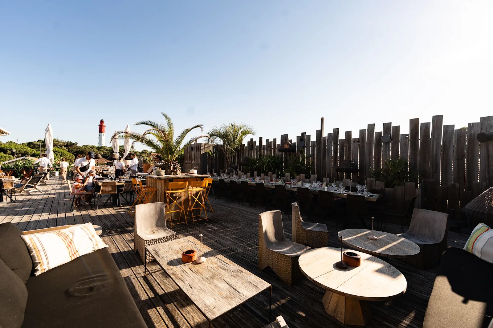 Terrasse en bois de restaurant en plein air avec chaises, tables dressées, plantes en pots et un phare blanc et rouge en arrière-plan sous un ciel bleu clair.