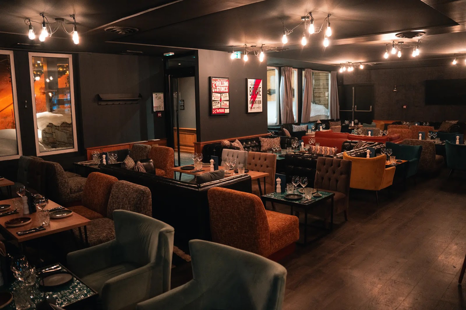 Intérieur chaleureux et moderne d'un restaurant avec des fauteuils et canapés colorés disposés autour de petites tables dressées de verres, assiettes et couverts, éclairage tamisé avec des ampoules suspendues et deux affiches musicales sur un mur sombre.