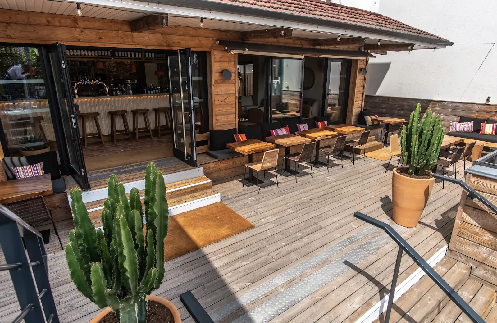Terrasse extérieure en bois avec tables, chaises et grands cactus en pots, devant l'entrée d'un bar avec porte-fenêtres ouvertes et tabourets au comptoir à l'intérieur.
