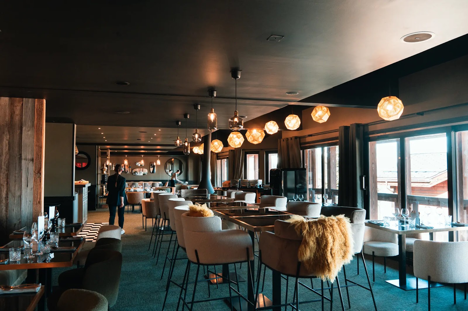 Intérieur élégant et chaleureux d'un restaurant moderne avec des tables équipées de chaises beige et marron, un éclairage suspendu design, des grandes fenêtres donnant sur l'extérieur et deux serveurs en train de préparer le lieu.