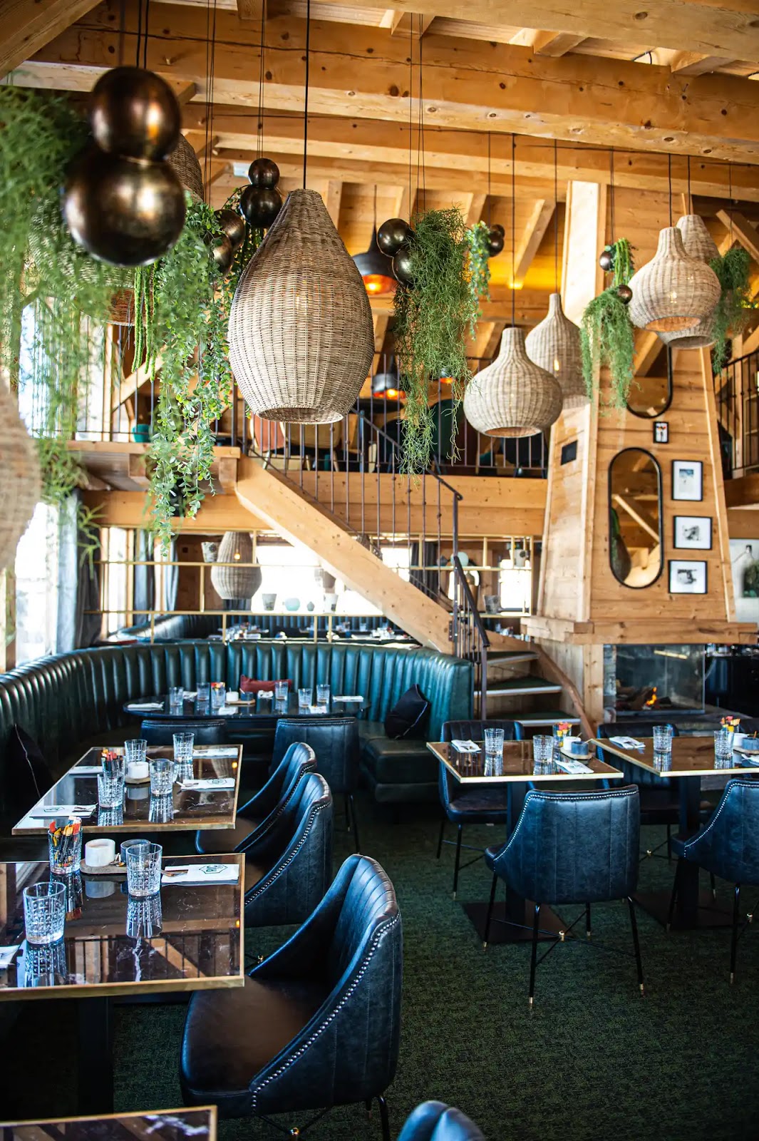 Intérieur de restaurant moderne avec murs et plafond en bois, grandes suspensions en osier, plantes suspendues, fauteuils en cuir noir autour de tables en marbre, et une cheminée encadrée de cadres photos.