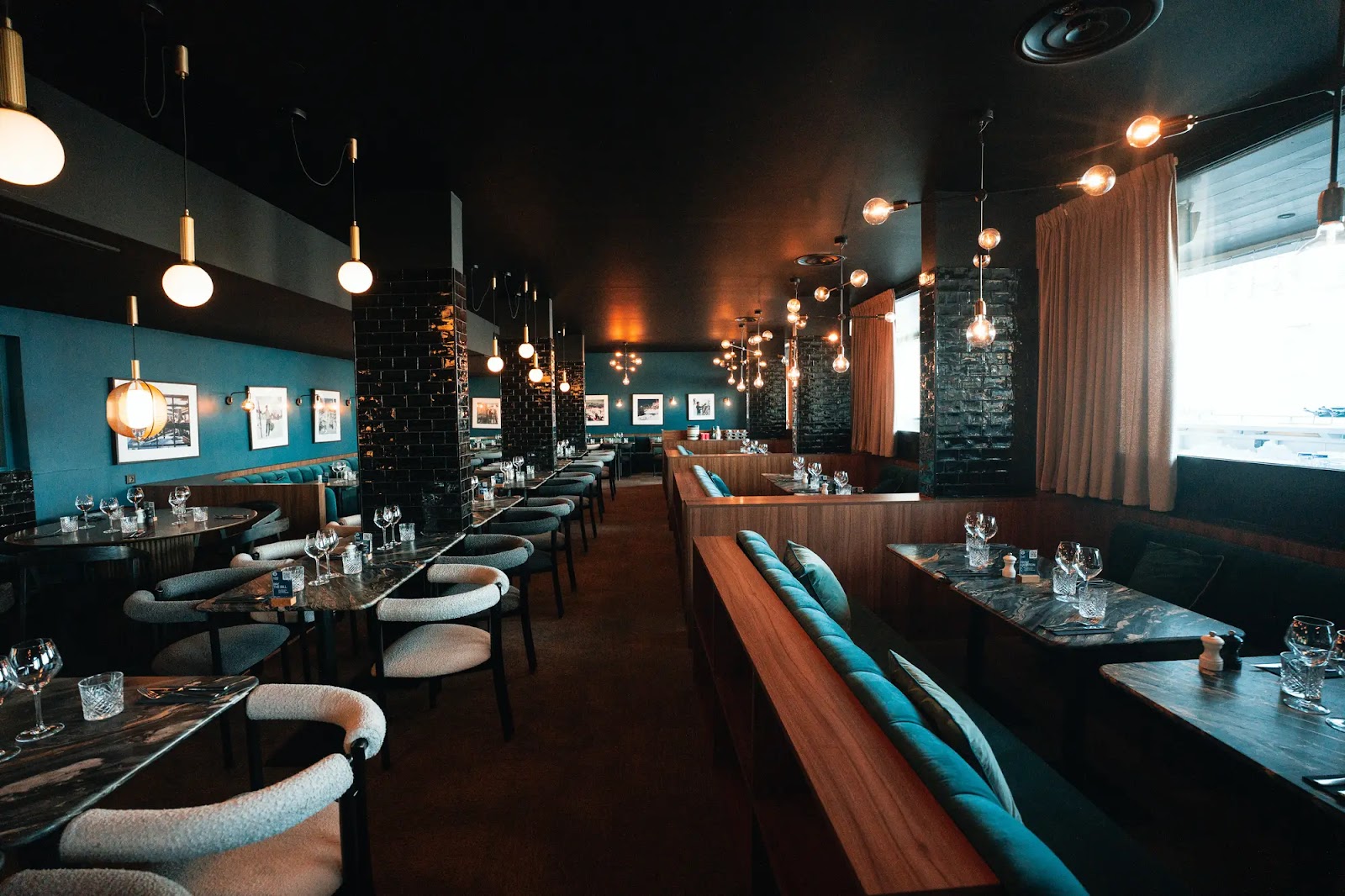 Intérieur d'un restaurant moderne avec des tables en marbre, chaises confortables et banquettes en bois avec coussins bleus, éclairage suspendu chaleureux et murs décorés de photos encadrées.