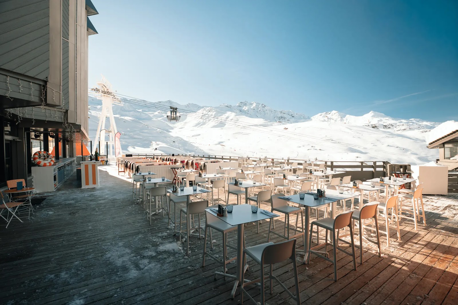 Terrasse de restaurant avec tables et chaises blanches sur un sol en bois, au pied de montagnes enneigées sous un ciel bleu clair, avec une télécabine visible en arrière-plan.