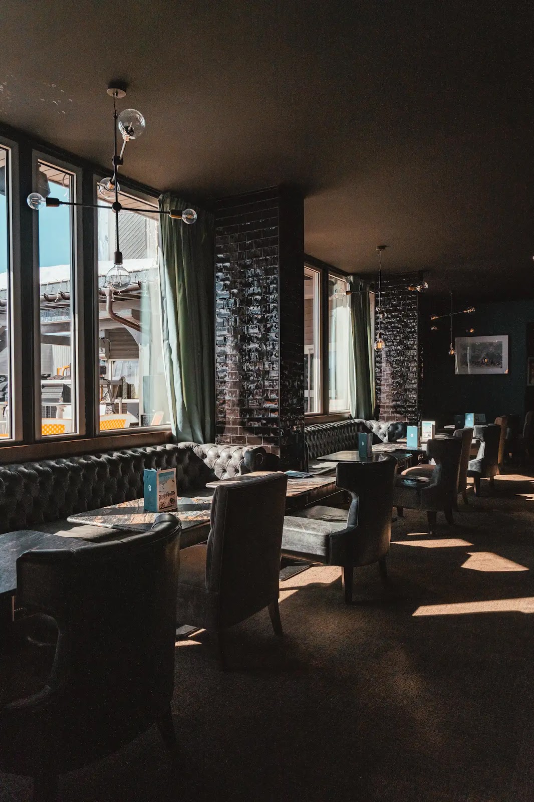 Intérieur sombre d'un café ou restaurant avec fauteuils en cuir, banquettes capitonnées, tables en pierre, murs en briques noires, grandes fenêtres et rideaux verts, lumière naturelle créant des ombres.