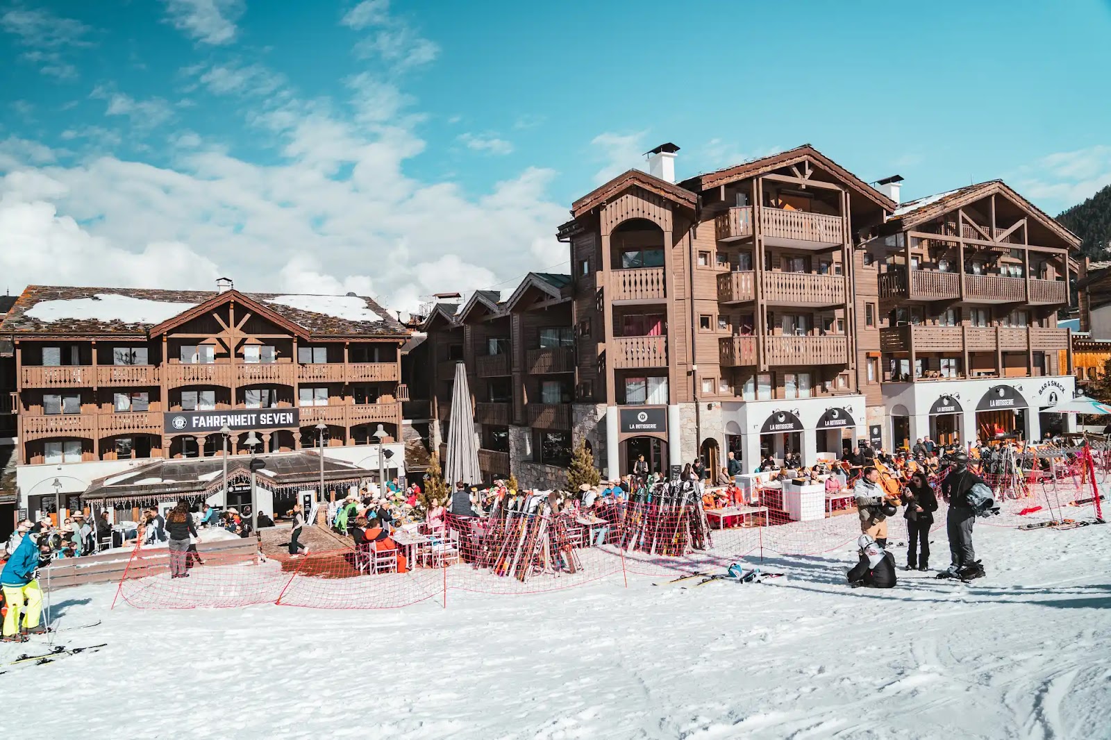 Vue d'un centre de ski animé avec des personnes en tenue de ski sur la neige près de bâtiments en bois comprenant un restaurant nommé Fahrenheit Seven et une rotisserie, sous un ciel bleu avec quelques nuages.