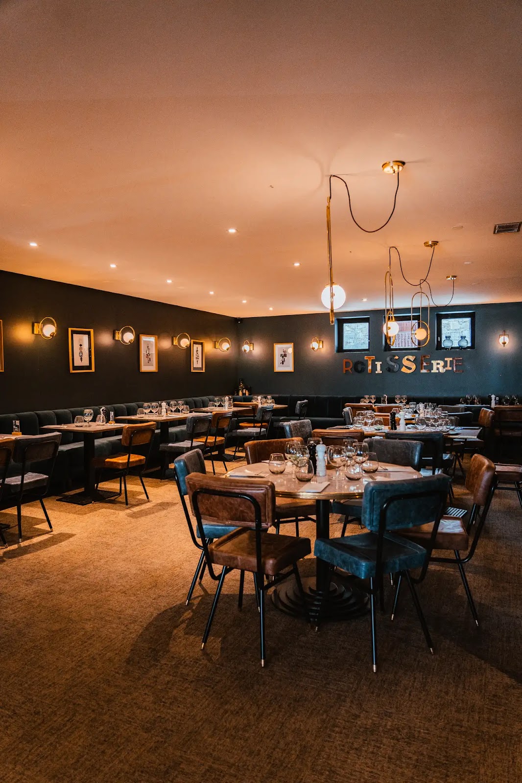 Intérieur d'un restaurant élégant avec des tables en bois rondes et rectangulaires, chaises en cuir marron et bleu, murs gris foncé décorés de cadres et d'appliques lumineuses, luminaires suspendus modernes, et sur un mur l'inscription 'ROTISSERIE'.