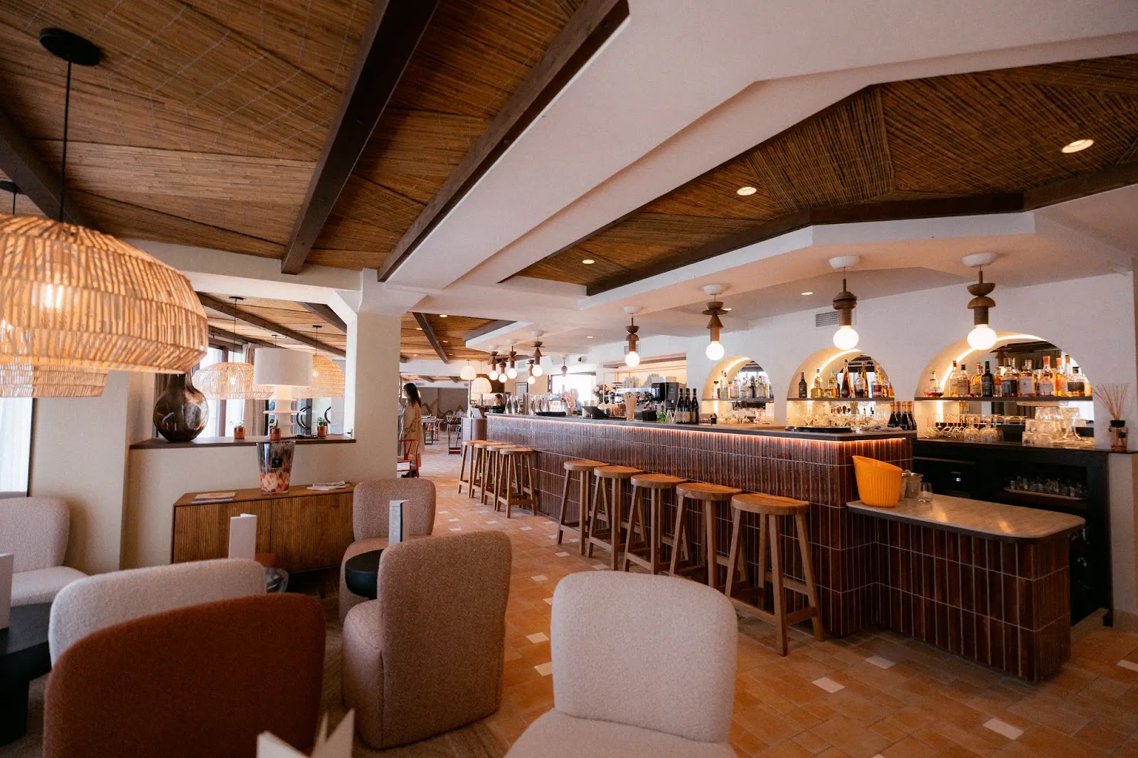 Intérieur élégant d'un bar avec un long comptoir en bois sombre muni de tabourets en bois, de nombreuses bouteilles et verres sur des étagères éclairées par des lampes suspendues rondes. Des fauteuils beige et marron sont disposés dans l'espace de détente, avec un plafond en bois et plusieurs luminaires en osier suspendus au-dessus.
