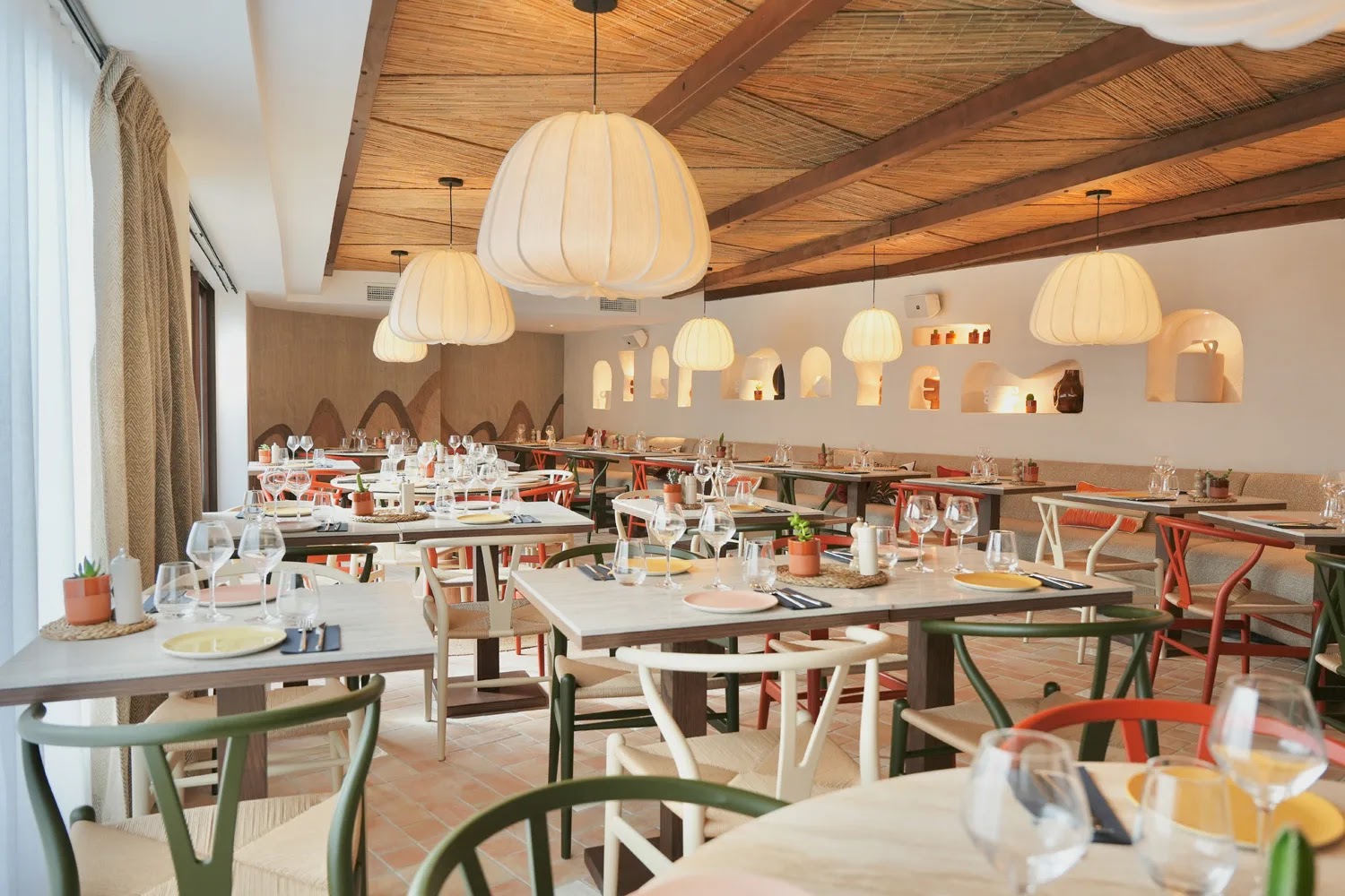 Salle de restaurant moderne avec tables en bois clair dressées de verres, assiettes colorées et petits pots de plantes, chaises variées vertes, blanches et rouges, lampes suspendues rondes en tissu blanc et plafond en bois avec nattes tressées.