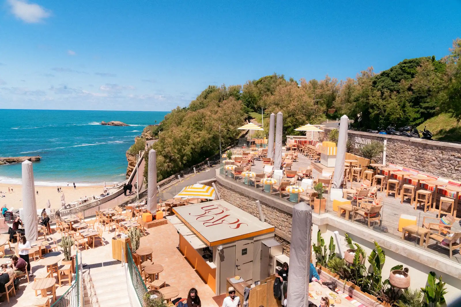 Terrasse de restaurant en plein air avec mobilier en bois et parasols jaunes et blancs, surplombant une plage de sable et une mer turquoise sous un ciel bleu clair, avec des arbres verts à l'arrière.