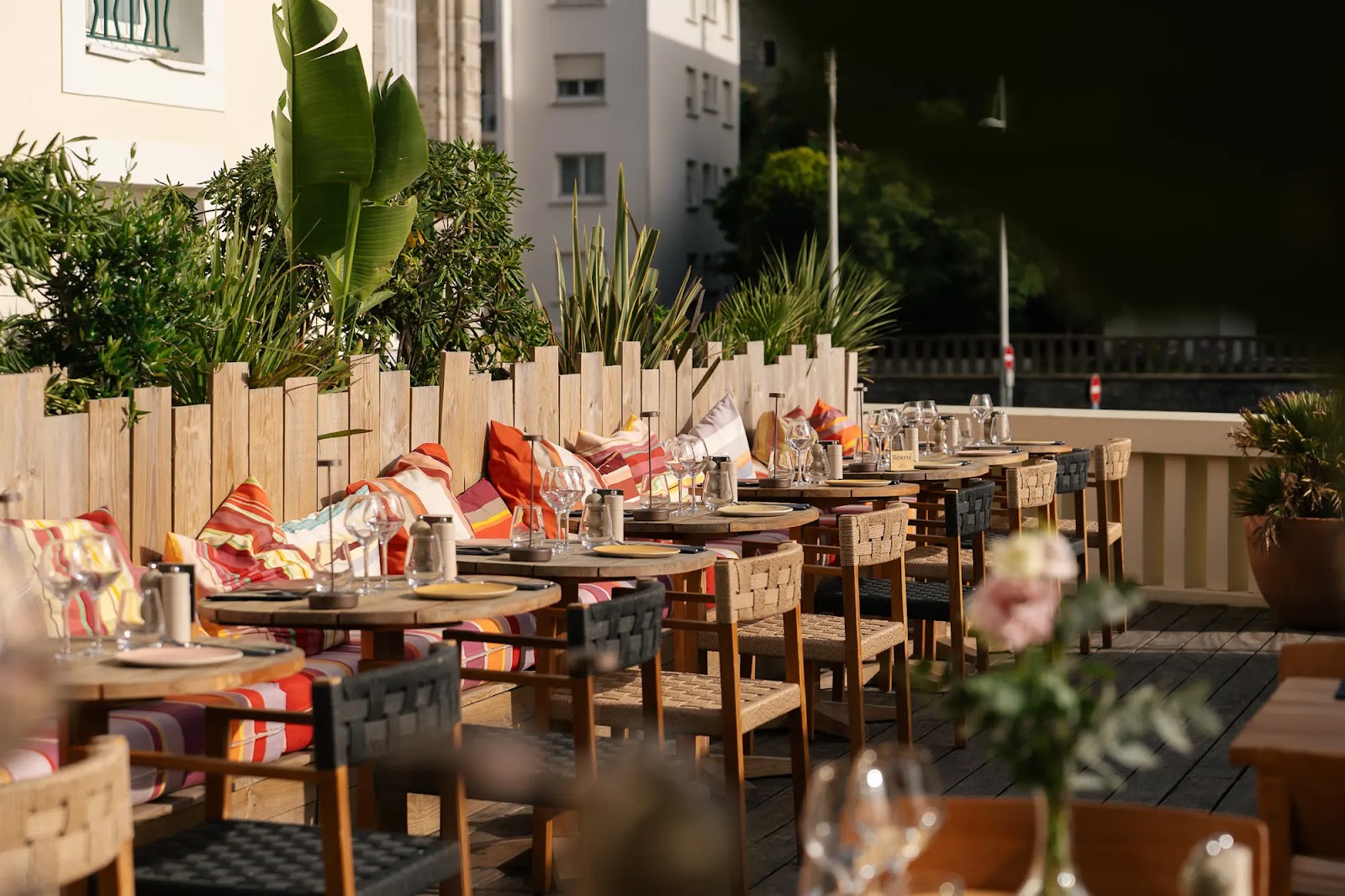 Terrasse de restaurant extérieure avec tables en bois circulaires, chaises en bois tressées, et banquette garnie de coussins colorés rayés rouge, orange et blanc, décorée de verres à vin et assiettes, entourée de plantes en pots et feuillages verts.