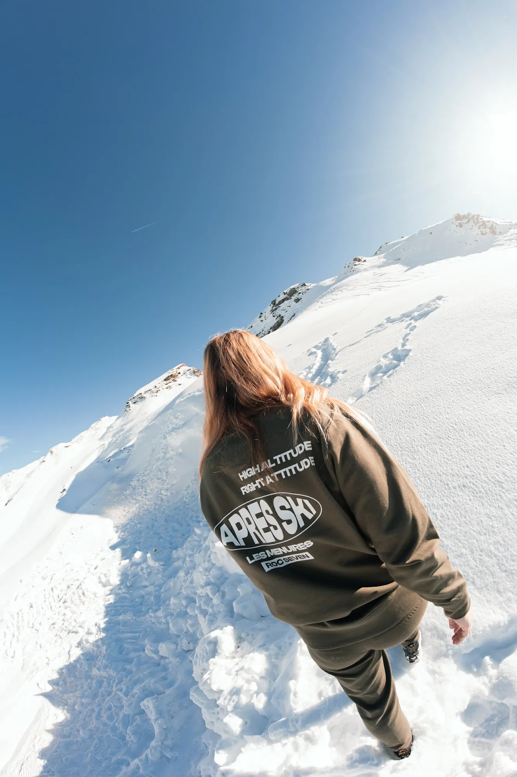 Personne vue de dos marchant sur un sentier enneigé en montagne par une journée ensoleillée avec un ciel bleu clair; la personne porte un ensemble kaki avec l'inscription « APRES SKI LES MENUIRES » dans le dos.