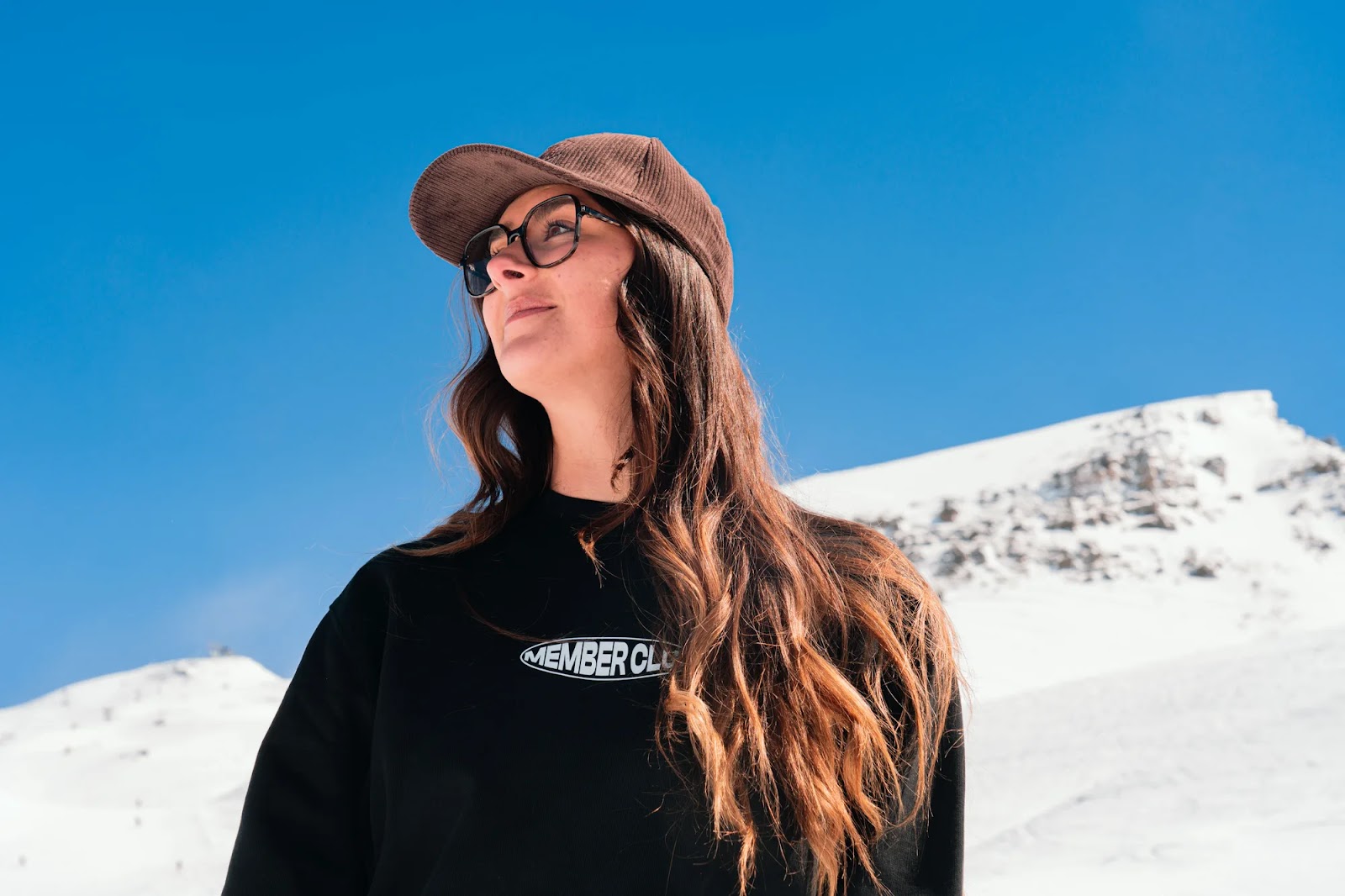 Jeune femme portant une casquette marron et des lunettes de soleil noires, vêtue d'un sweat-shirt noir avec l'inscription MEMBER CLUB, en extérieur avec un ciel bleu clair et une montagne enneigée en arrière-plan.