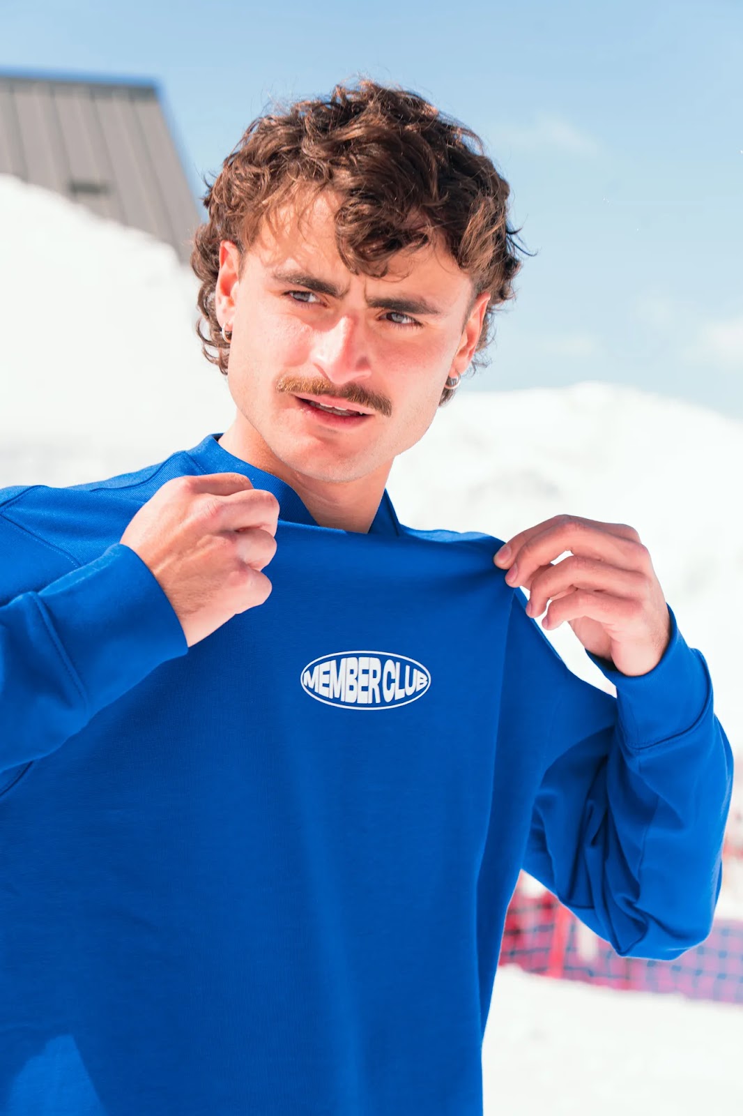 Homme aux cheveux bouclés et moustache portant un sweat bleu vif avec l'inscription "MEMBER CLUB" en blanc sur la poitrine, tenant le tissu près du cou, en extérieur sous un ciel clair avec un fond enneigé.
