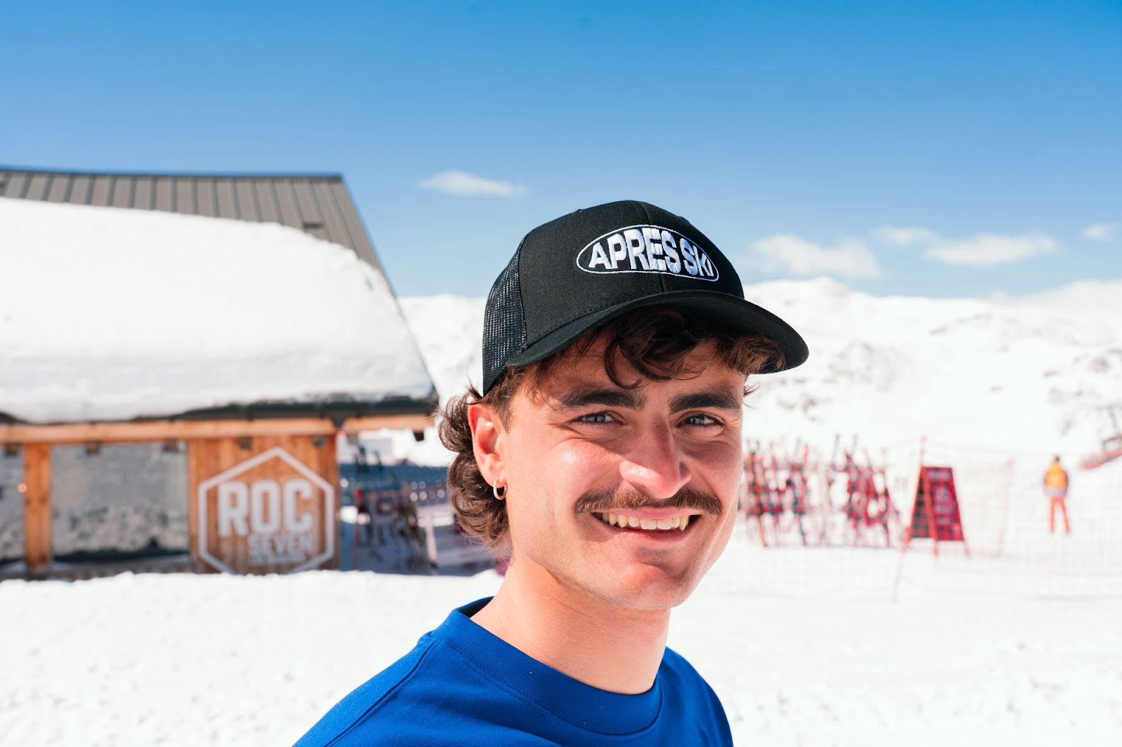Jeune homme souriant portant une casquette noire avec l'inscription «APRÈS SKI» devant un paysage enneigé avec un bâtiment en bois et un ciel bleu clair.