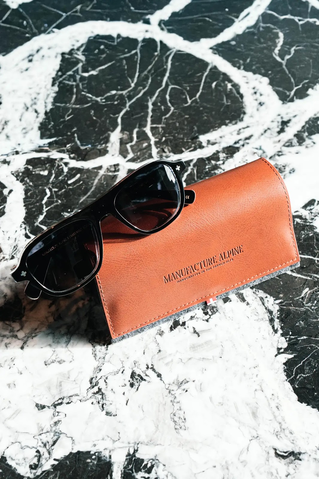 Lunettes de soleil noires posées sur un étui à lunettes en cuir marron sur une surface en marbre noir et blanc, l'étui porte l'inscription Manufacture Alpine.