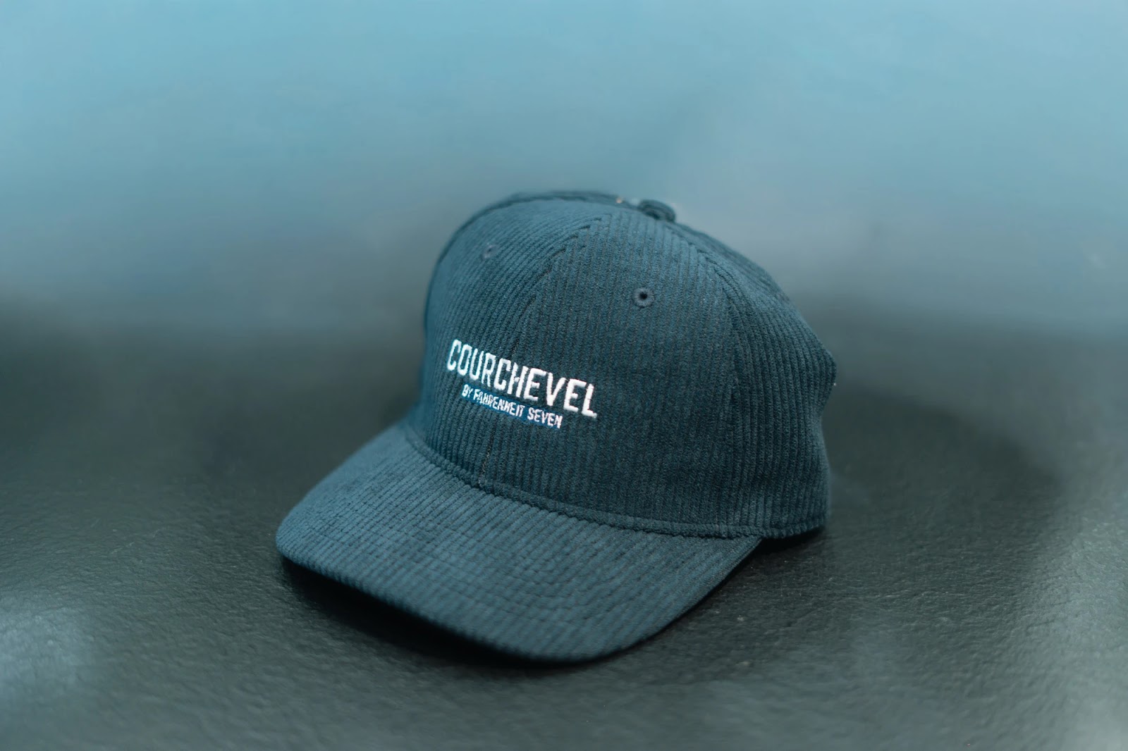 Casquette bleu foncé en velours côtelé posée sur une surface noire, avec le texte brodé blanc « COURCHEVEL BY FAHRENHEIT SEVEN » sur le devant.