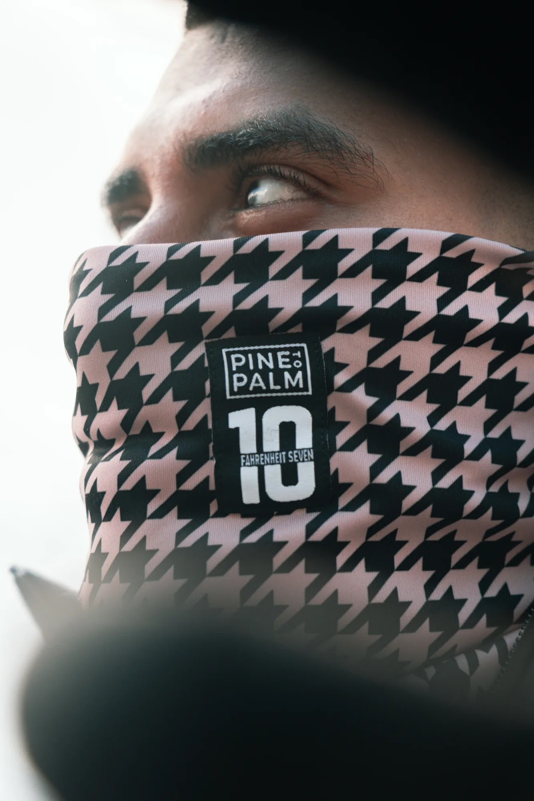 Gros plan sur le visage d'un homme portant un masque couvrant son nez et sa bouche, avec un motif pied-de-poule noir et rose et une étiquette avec le texte "PINE PALM 10 FAHRENHEIT SEVEN".