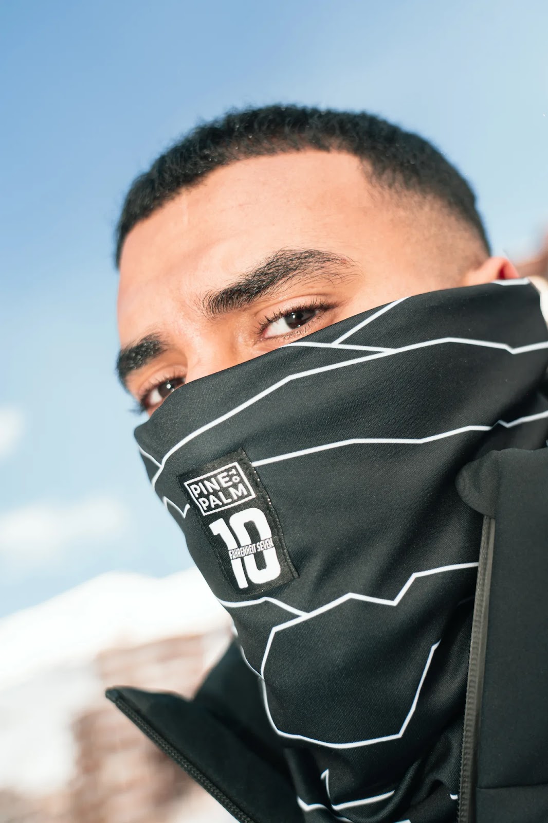 Portrait rapproché d'un homme portant un masque noir avec des lignes blanches et un logo indiquant "Pine & Palm 10 Fahrenheit Seven", avec un ciel bleu en arrière-plan.