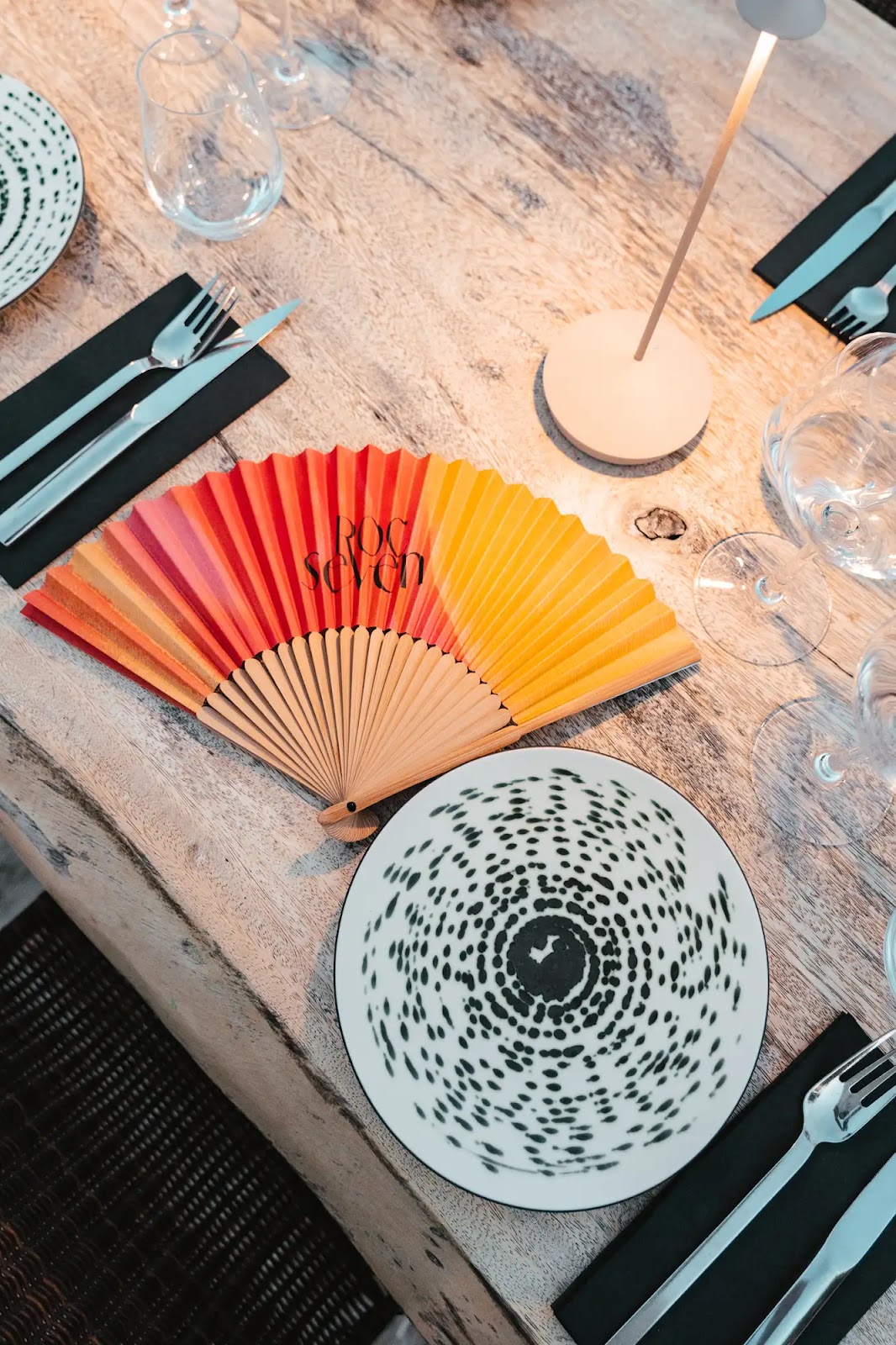 Table en bois clair dressée avec des assiettes blanches à motifs noirs, des verres à vin, des couverts sur des serviettes noires et un éventail déployé aux couleurs dégradées rouges, oranges et jaunes avec le texte « ROC seven ».
