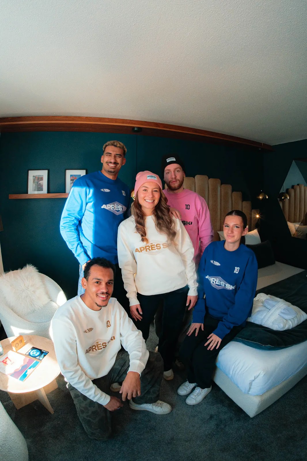 Cinq personnes portent des pulls colorés avec le logo 'APRÈS SKI' dans une chambre moderne avec murs foncés, fauteuil blanc et lit avec peignoir. Trois debout, deux assis, tous souriants. Une femme au centre porte un bonnet rose, un homme à l'arrière un bonnet noir, les autres ont des pulls bleu ou crème. Atmosphère chaleureuse et conviviale.