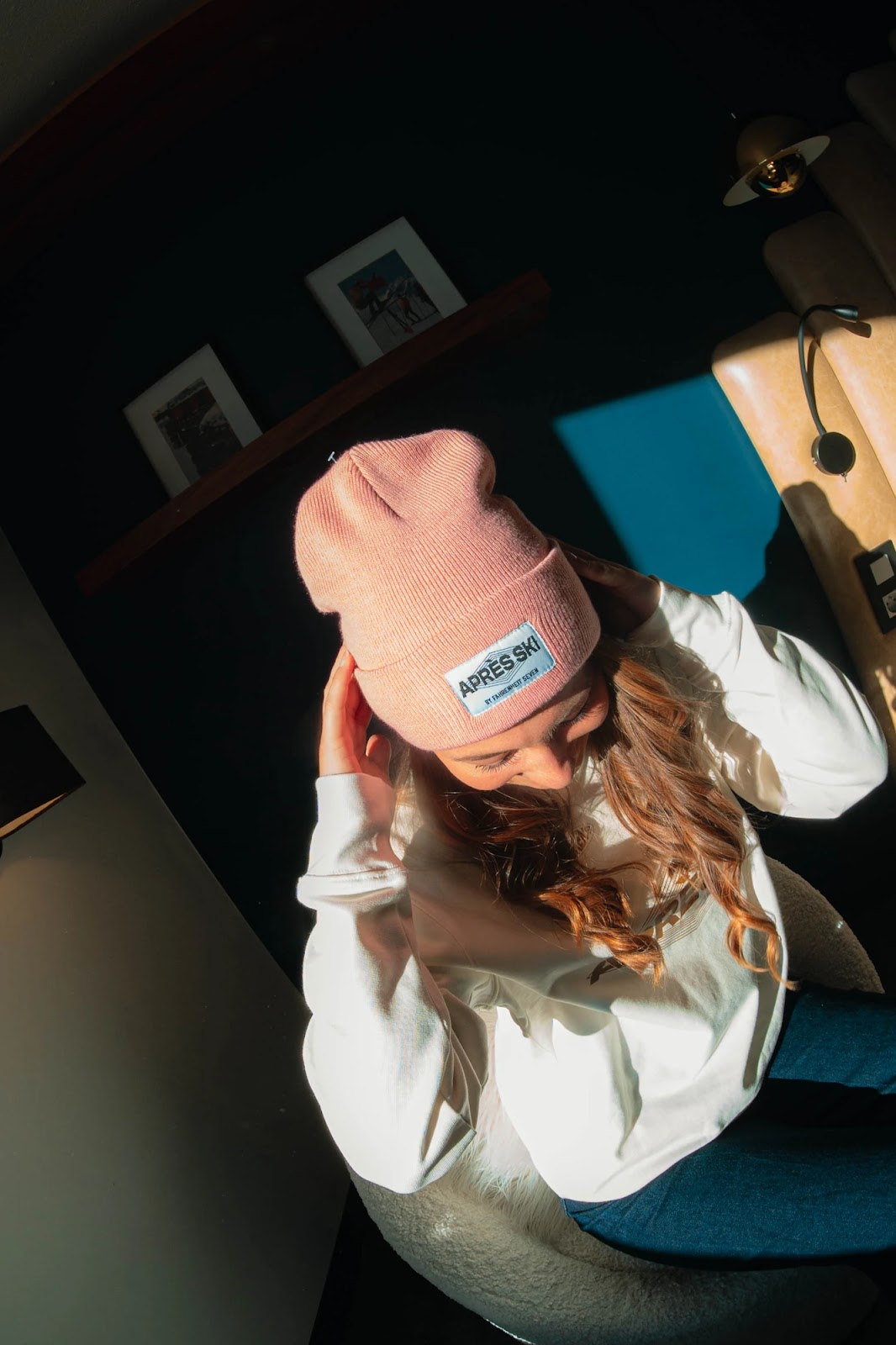 Jeune femme assise, portant un bonnet rose avec une étiquette "APRES SKI" et un pull blanc, éclairée par une lumière vive venant de la droite.