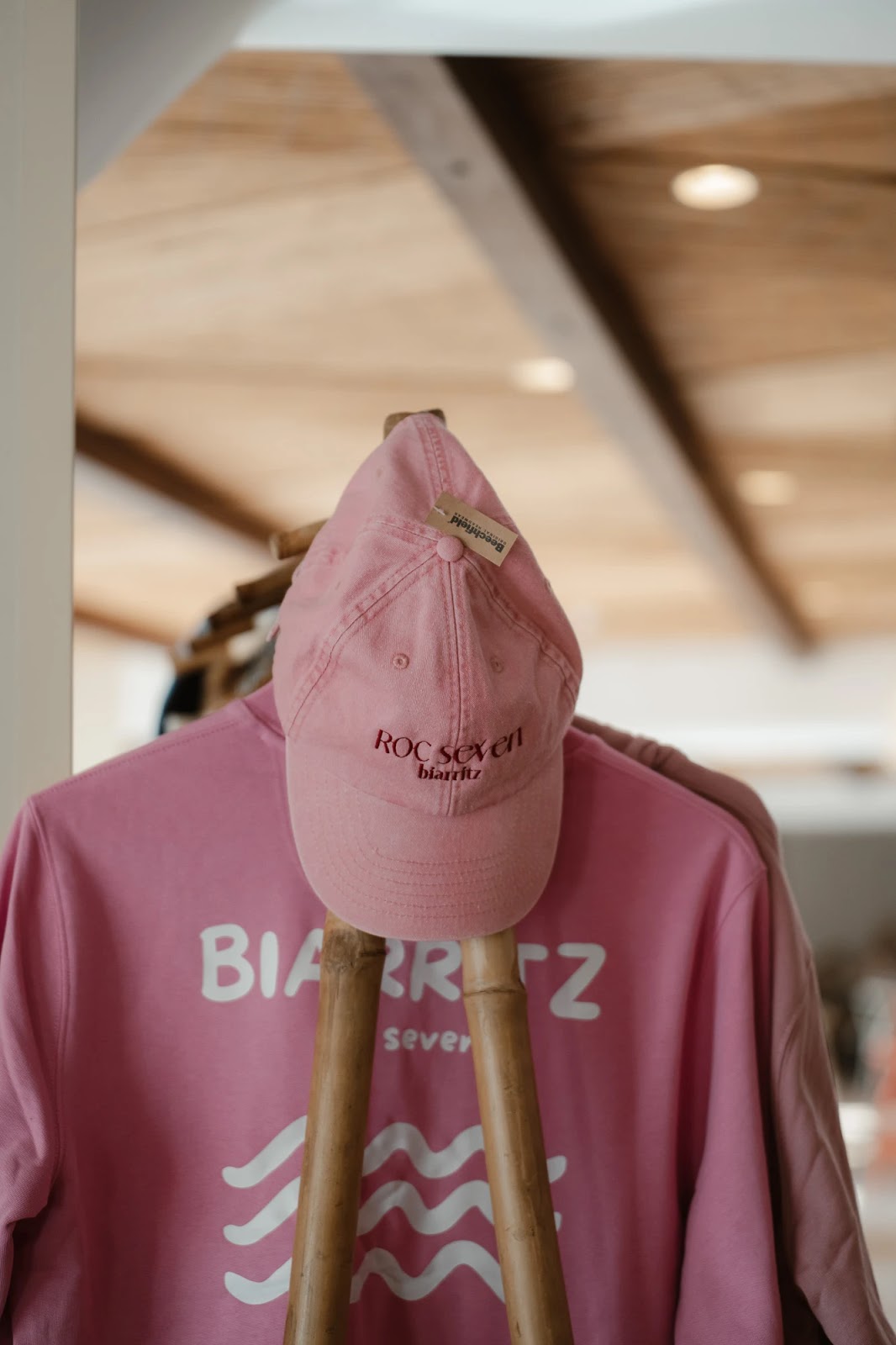 Casquette rose avec inscription "ROC seven biarritz" accrochée sur un vêtement rose portant l'inscription "BIARRITZ" en lettres blanches, suspendue sur un support en bois à l'intérieur d'un espace avec un plafond en bois.