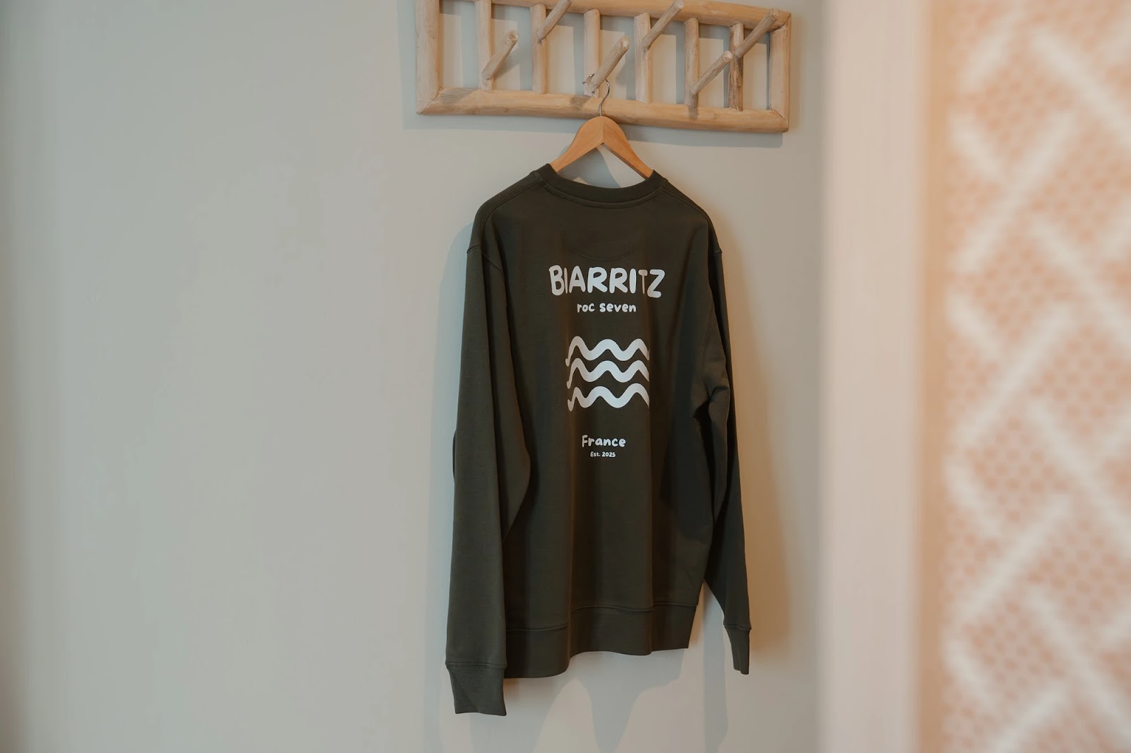 Pull kaki suspendu sur un cintre en bois contre un mur clair, avec le texte blanc « BIARRITZ roc seven France Est. 2025 » et un dessin de vagues.