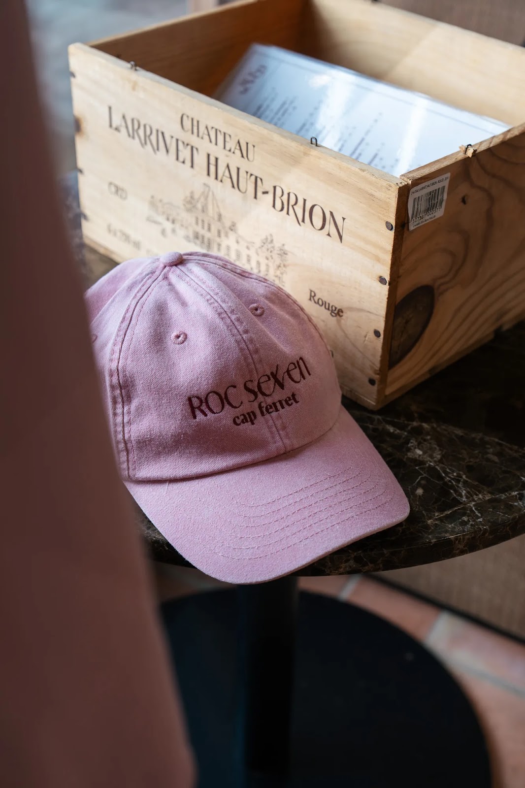 Casquette rose avec inscription "ROC sex en cap ferret" posée sur une table en marbre devant une caisse en bois marquée "CHATEAU LARRIVET HAUT-BRION".