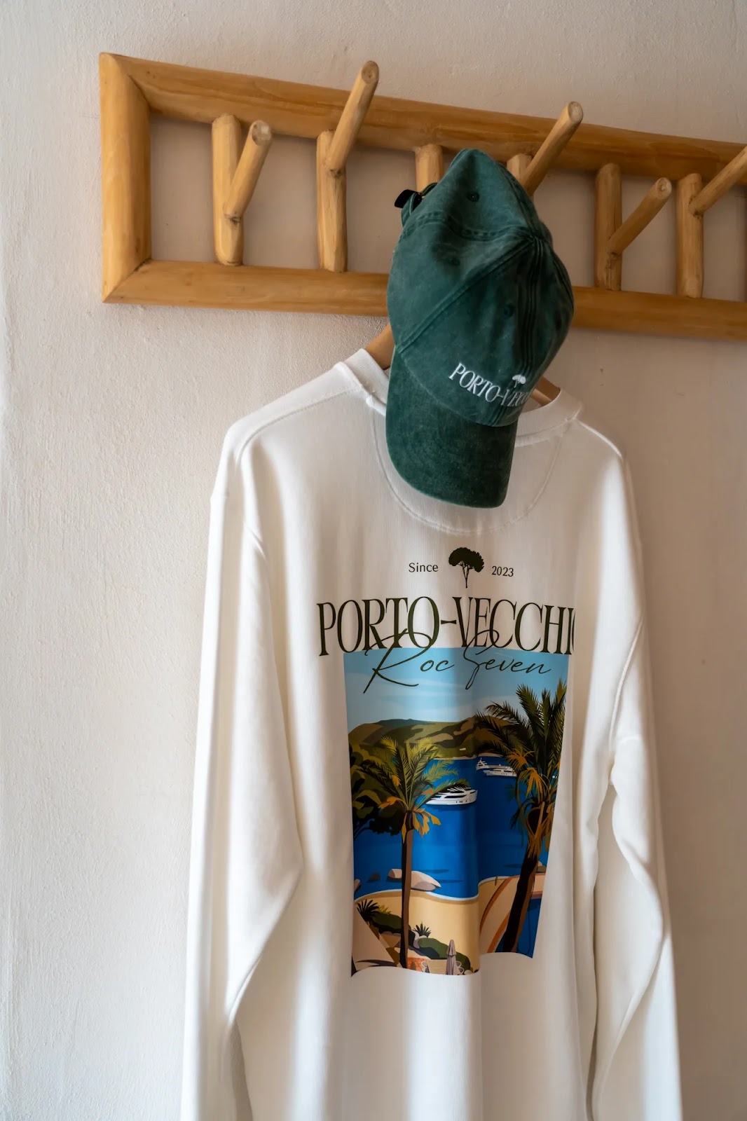 Un sweat-shirt blanc accroché sur un cintre sous un porte-manteau en bois, avec une casquette verte posée sur le cintre. Le sweat-shirt affiche une illustration colorée d'une baie avec des palmiers, des bateaux et des collines, accompagnée du texte « Porto-Vecchio Roc Seven » et « Since 2023 ». La casquette verte porte aussi le texte « Porto-Vecchio ».
