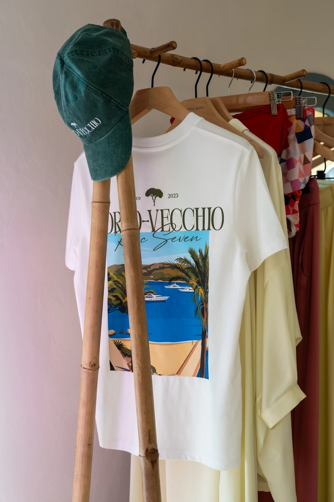 Vêtements suspendus sur un portant en bois, avec un t-shirt blanc affichant une illustration d'un paysage marin avec des yachts, des palmiers et du sable, et une casquette verte accrochée au portant.