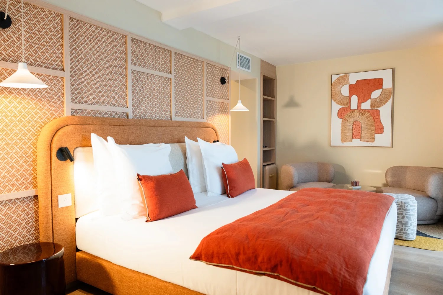 Chambre d'hôtel moderne avec un grand lit double aux coussins blancs et deux coussins décoratifs orange, une couverture orange, un mur avec un panneau en osier, deux suspensions blanches au-dessus du lit, une table d'appoint ronde marron, deux fauteuils beiges disposés autour d'une petite table basse, et un tableau abstrait aux couleurs orangées et beiges sur un mur beige.