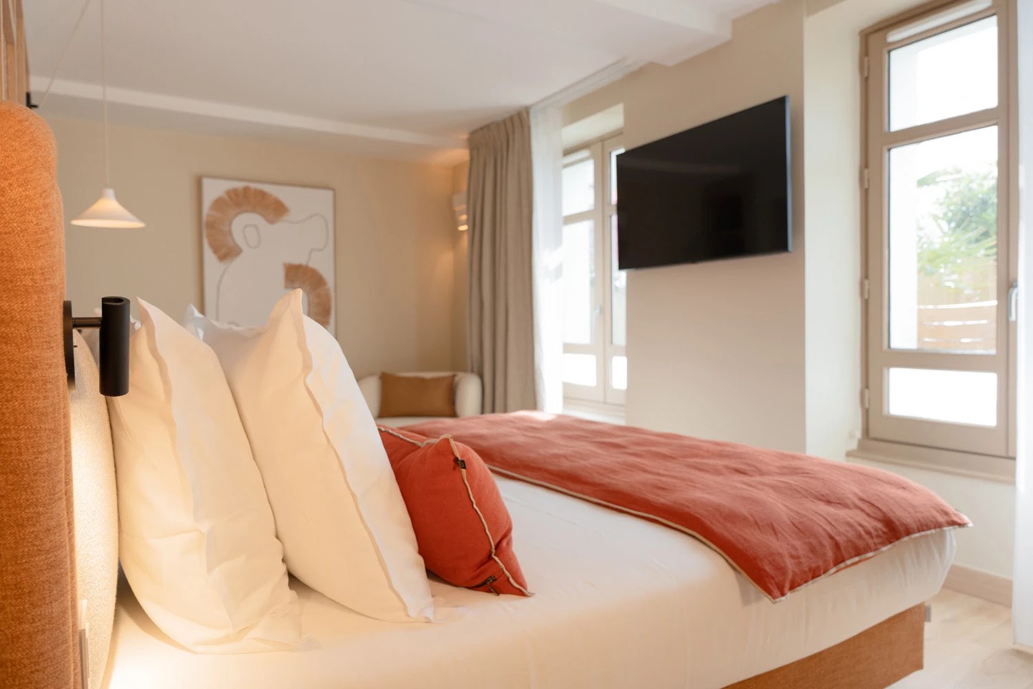 Chambre moderne avec un grand lit aux draps blancs et une couverture orange, deux oreillers blancs et un coussin orange. Un fauteuil beige est visible dans le coin, un tableau abstrait est accroché au mur beige, un téléviseur est fixé au mur à côté de grandes fenêtres, laissant entrer la lumière naturelle.