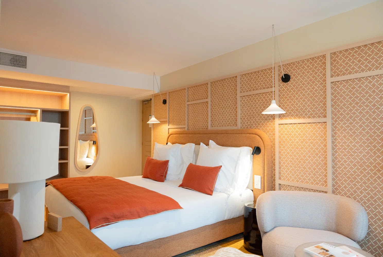 Chambre d'hôtel moderne avec un grand lit double habillé de draps blancs, coussins et jeté de lit orange, deux lampes suspendues blanches au-dessus, un mur décoratif en tissage beige, une chaise beige et une table avec des magazines.