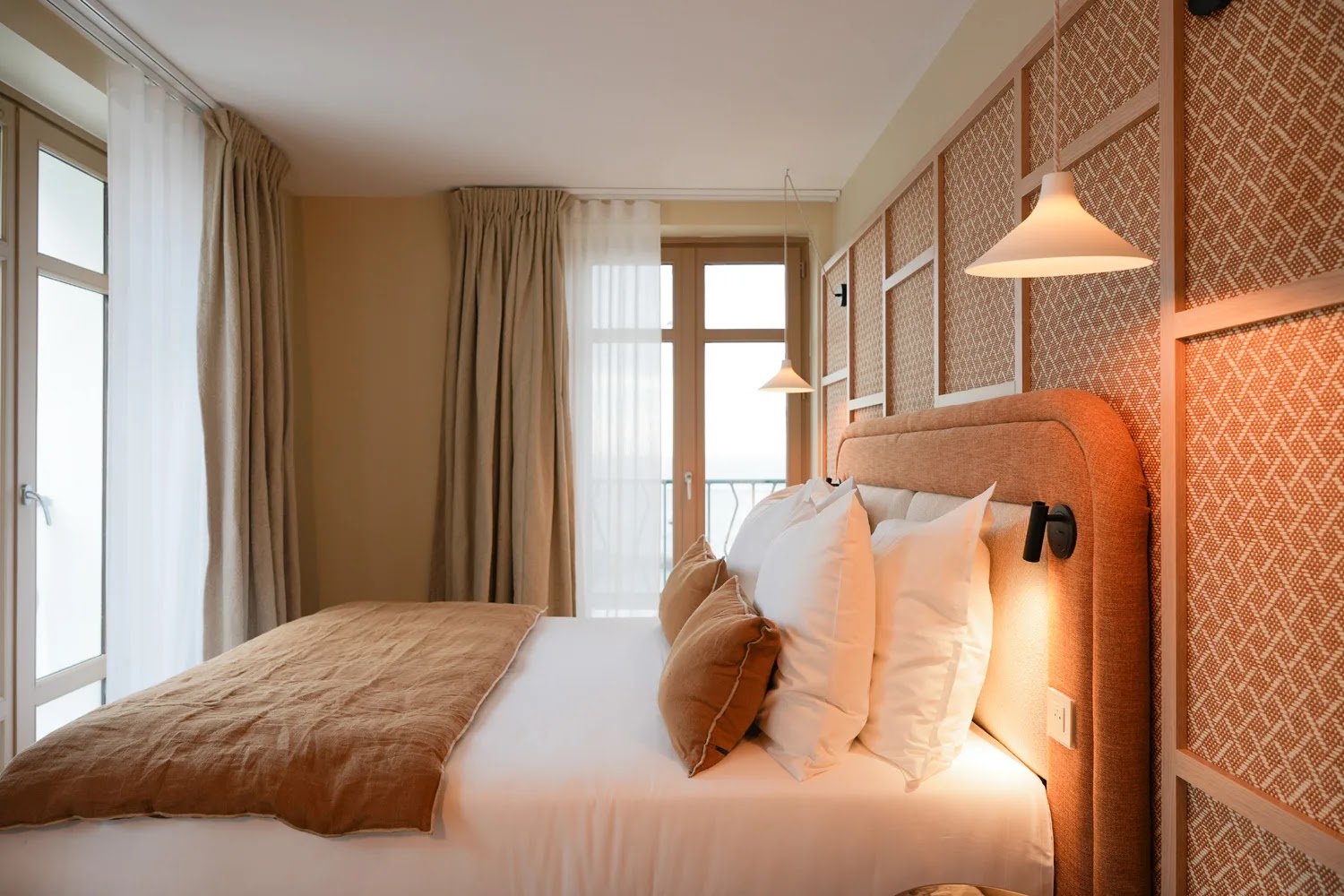 Chambre à coucher lumineuse avec lit double, couverture marron, plusieurs oreillers blancs et marron, têtes de lit rembourrées, fenêtres avec rideaux beige, et lampes suspendues au plafond.